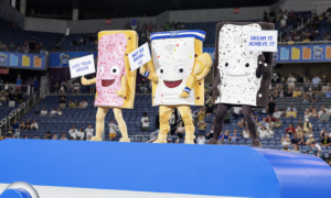 Three cartoon dessert mascots stand on a blue stage in a stadium, each holding a sign with motivational messages: 'Live Your Dream', 'Not My Dream... Yet', and 'Dream It, Achieve It'