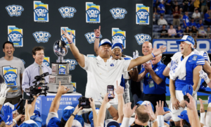 Team celebrates victory on stage with a large trophy at the Pop-Tarts Bowl, fans cheering in the foreground.