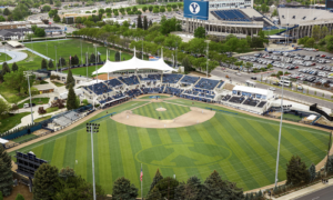Aerial view of a baseball stadium with a striped infield, surrounding blue seating, and a large blue BYU scoreboard in the background.
