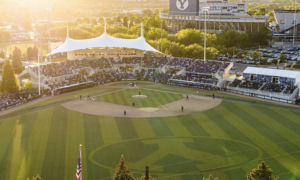 Overhead view of a baseball stadium at sunset with a crowded grandstand and a white canopied structure over the press box area on the leftglow.