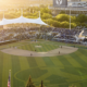 Overhead view of a baseball stadium at sunset with a crowded grandstand and a white canopied structure over the press box area on the leftglow.