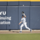 Baseball player in a white uniform reaches high to catch a ball against a blue wall that reads 'BYU Continuing Education'.