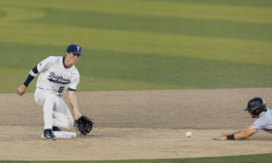 Baseball infielder in a Brighton uniform guards second base, glove ready as a runner slides in and the ball rolls toward him.