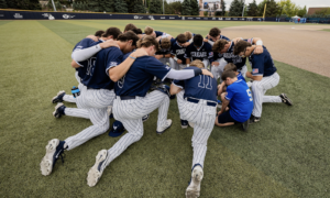 Baseball team in navy jerseys with white pinstripes bow in a huddle on the infield, arms around each other in a pre-game or motivational moment on the field.