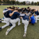 Baseball team in navy jerseys with white pinstripes bow in a huddle on the infield, arms around each other in a pre-game or motivational moment on the field.
