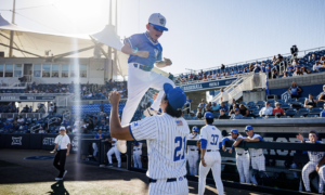 A young baseball player in blue jersey and white cap jumps onto a teammate in pinstriped uniform near the dugout as fans watch from the stands.