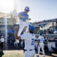 A young baseball player in blue jersey and white cap jumps onto a teammate in pinstriped uniform near the dugout as fans watch from the stands.