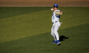 Baseball pitcher in white pinstripe uniform throws a ball on the field, wearing blue cap and sunglasses.