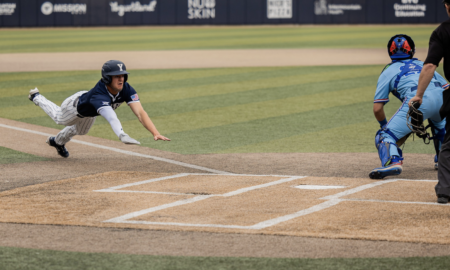 Baseball player in navy uniform slides into home plate as the catcher in blue gear closes in from behind with his glove ready.