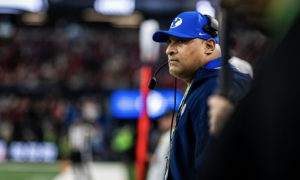 Side profile of a football coach wearing a blue BYU cap and headset, standing on the sideline during a game with a blurred crowd in the background, focused on the field.