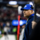 Side profile of a football coach wearing a blue BYU cap and headset, standing on the sideline during a game with a blurred crowd in the background, focused on the field.