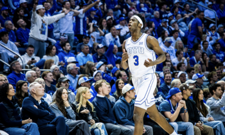BYU basketball player wearing number 3 in a white uniform runs down the court as a packed crowd in blue cheers in the background.
