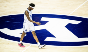 Basketball player in a white jersey with blue trim (number 3) walks across a wooden court over a large blue-and-white logo.