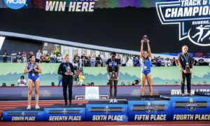 Five athletes stand on a multi-tier awards podium at an NCAA track and field championships ceremony, proudly displaying medals and trophies against a large “Champions Win Here” banner.