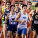 Group of male track runners in a race on an outdoor track, wearing colorful uniforms and race bibs. Ahead are BYU and W outfits, with intense expressions.