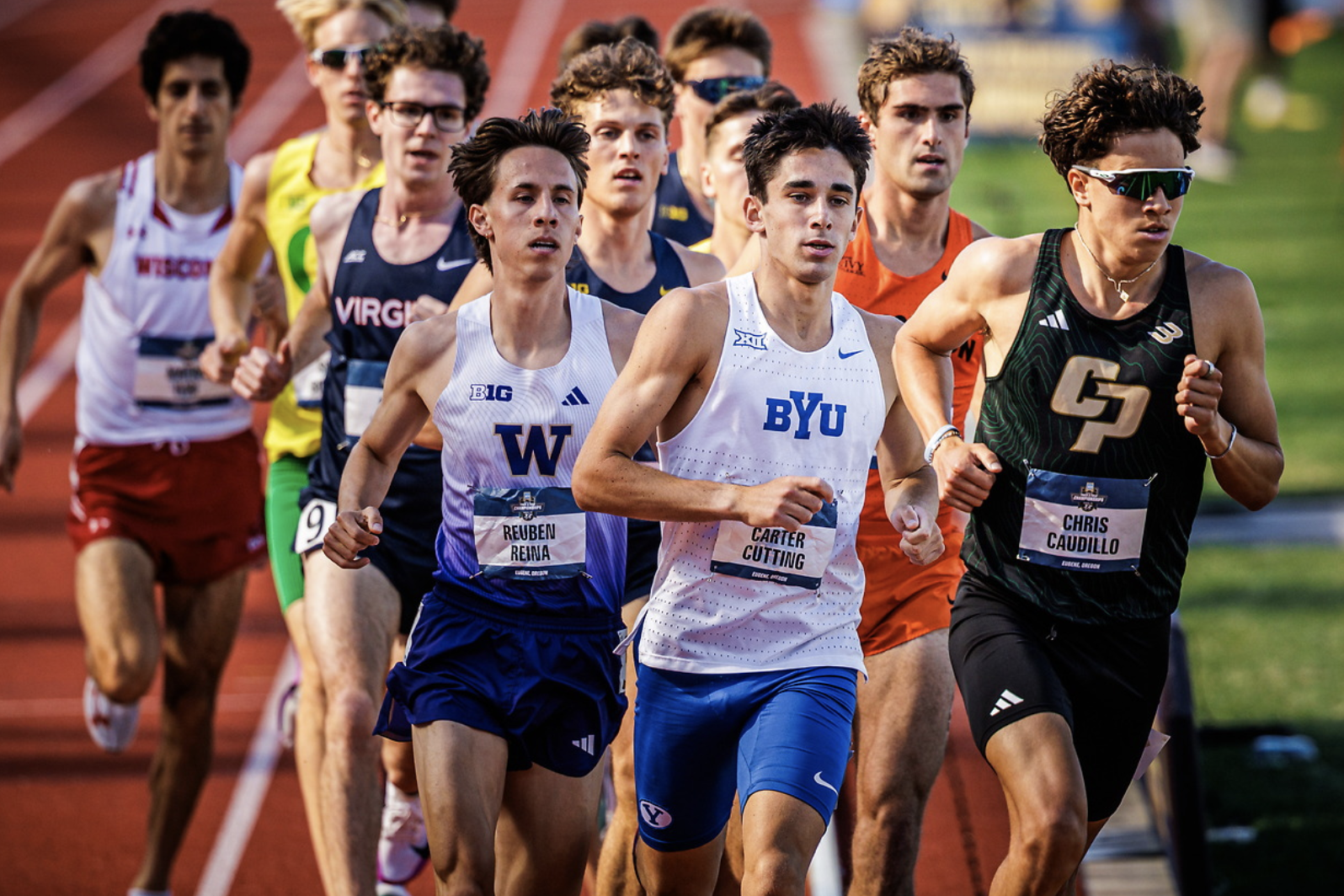 Group of male track runners in a race on an outdoor track, wearing colorful uniforms and race bibs. Ahead are BYU and W outfits, with intense expressions.