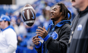 A man in a blue-hooded jacket watches a football spinning toward him in midair at a stadium filled with blue-clad fans.