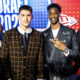 Two young men pose on a bright red/blue stage with a 'DRAFT' backdrop, wearing stylish jackets; one shows a peace sign.