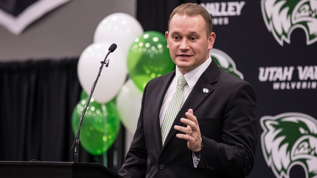 Man in a dark suit and light green striped tie speaks at a podium with a microphone, backed by Utah Valley Wolverines branding and green/white balloons.
