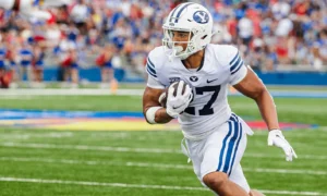 BYU football player in white uniform running with ball during a game with a crowd in the stands behind him