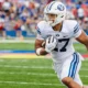 BYU football player in white uniform running with ball during a game with a crowd in the stands behind him