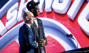 Two men in suits shake hands on a stage with a red, white, and blue backdrop and two microphones nearby, smiling at the event.