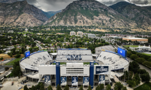 Aerial view of Lavell Edwards Stadium at BYU, with the football field and stands visible and a rugged mountain range in the distance behind the city.