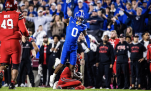 Blue-uniformed football player leaps over a tackling opponent on a packed sideline during a game, crowd cheering in the background, night scene