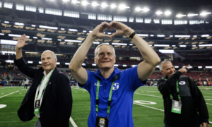 Three men on a football field celebrate in a stadium, with the center man forming a heart shape with his hands and smiling while two others wave nearby.