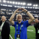 Three men on a football field celebrate in a stadium, with the center man forming a heart shape with his hands and smiling while two others wave nearby.