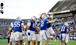 Football players in blue and white celebrate a touchdown on the field, with one player jumping in the air as teammates cheer nearby.