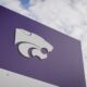 Close-up of a purple sign with a white abstract logo against a blue sky background.