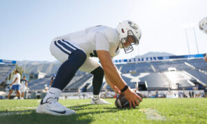 Football player in a white uniform and helmet crouches to touch a ball on the grass with the stadium in the background.