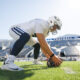 Football player in a white uniform and helmet crouches to touch a ball on the grass with the stadium in the background.