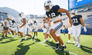 Group of BYU football players sprint onto a sunlit practice field in navy and white uniforms.
