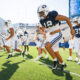 Group of BYU football players sprint onto a sunlit practice field in navy and white uniforms.