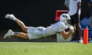 Football player in white uniform dives horizontally to catch a ball near an orange/red pylon on a sunny field.