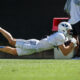 Football player in white uniform dives horizontally to catch a ball near an orange/red pylon on a sunny field.