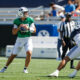 Quarterback in a green jersey prepares to throw a football during a game, with teammates and defenders nearby on the field.