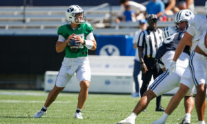 Quarterback in a green jersey holds the football, looking downfield as defenders close in during a sunny football game.