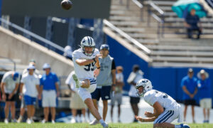 BYU football player in white jumps to catch an airborne ball as a teammate on the ground and spectators watch in the background.