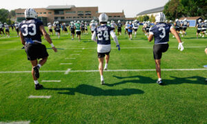Football players in navy uniforms run onto a practice field with a campus building in the background, during a drills session on green turf.
