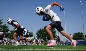 Football players sprinting on a sunny practice field; foreground player in white jersey and helmet with pink shoes ready to start.