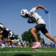 Football players sprinting on a sunny practice field; foreground player in white jersey and helmet with pink shoes ready to start.