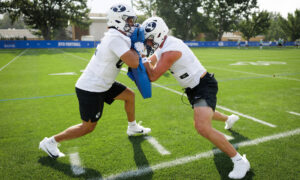 Two BYU football players in white jerseys collide during a practice drill on a sunny green field with a blue sideline banner behind them.”