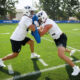 Two BYU football players in white jerseys collide during a practice drill on a sunny green field with a blue sideline banner behind them.”