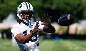 Football player in a white BYU helmet reaches for a football mid-air on a sunny practice field, white jersey.