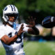 Football player in a white BYU helmet reaches for a football mid-air on a sunny practice field, white jersey.