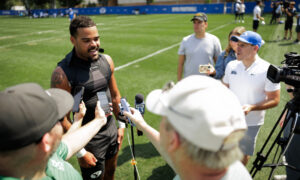Athlete in a black sleeveless shirt speaks to reporters on a grassy field, surrounded by microphones and cameras held by journalists and crew members.