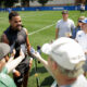 Athlete in a black sleeveless shirt speaks to reporters on a grassy field, surrounded by microphones and cameras held by journalists and crew members.
